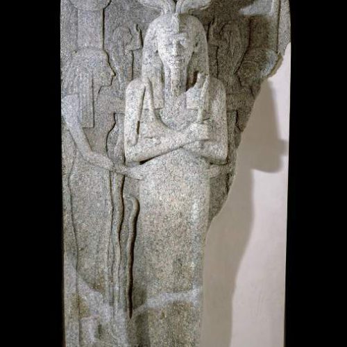 A large red granite lid of sarcophagus. There are three images of people carved into the granite, standing up. The central figure is male and protrudes from the lid, facing the viewer. His arms are crossed across his chest and he wears a beard, headpiece, and crown of feathers and horns. The two women either side of him face him and have their hands on his waist. The lid is broken on the right, and the side and the feet have been filled in in white plaster.