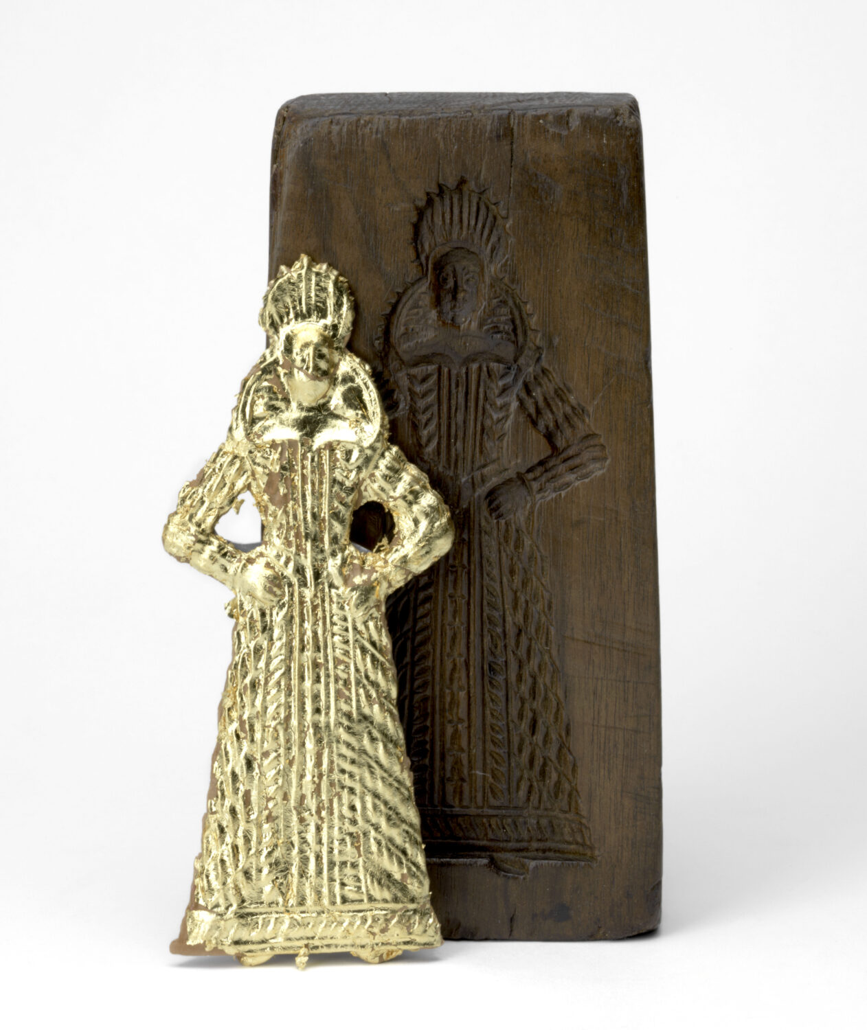 A gilded gingerbread woman stands upright in front of a the wooden mould from which she was baked.